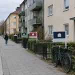 Lugn bostadsgata med till salu-skyltar på framsidan, en person går förbi, speglar bostadsmarknadens läge.