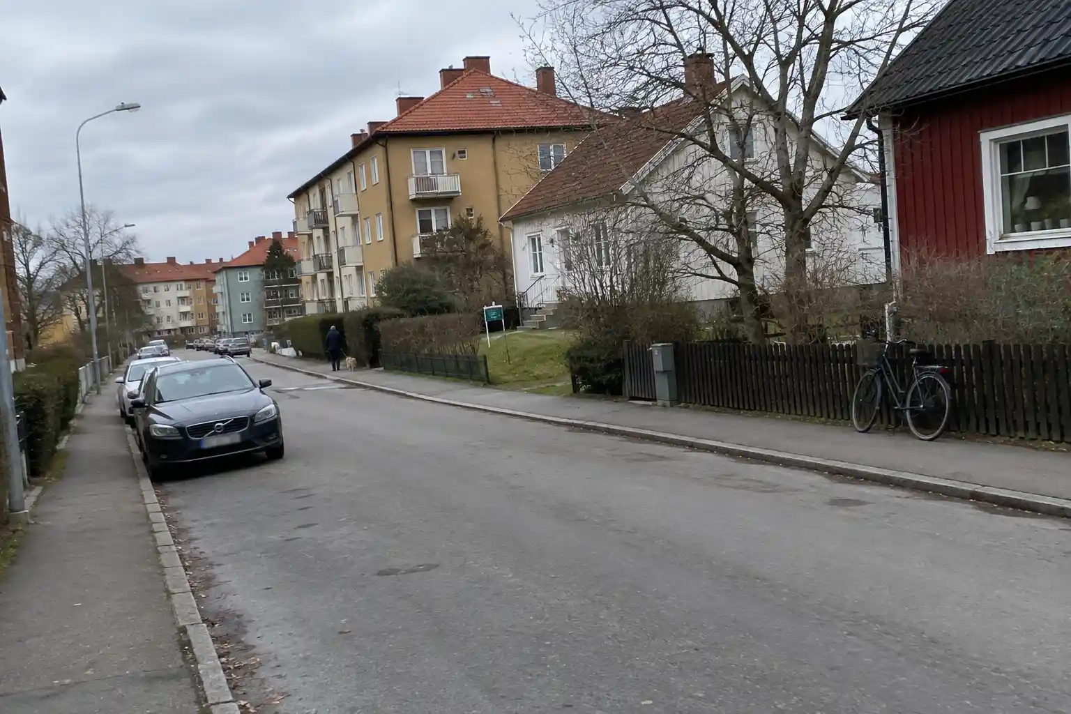 Bostadsgata i svenskt villaområde en mulen eftermiddag, person promenerar hund i bakgrunden, relevant för bostadsprisförvä...
