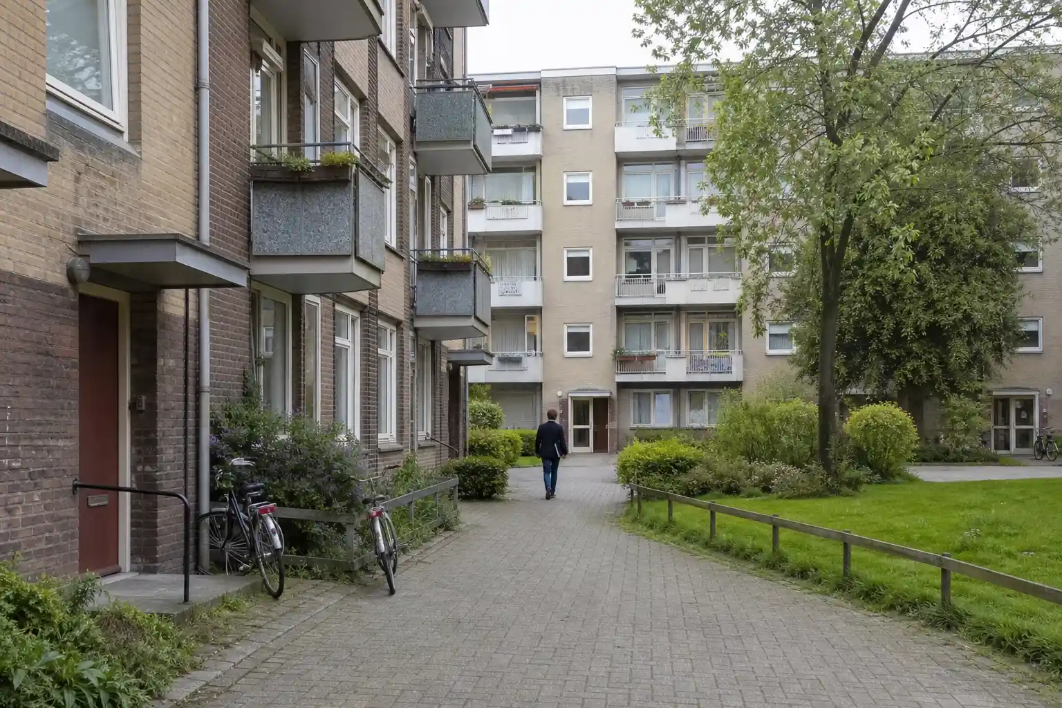 Bostadsgård mellan flerfamiljshus där en person går mot en entré, vardaglig miljö kopplad till fastighetsförvaltare uppköp